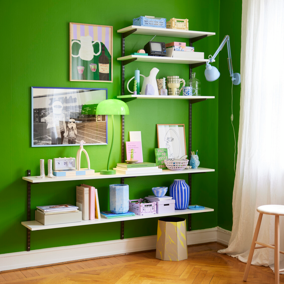 Genevieve sitter vid en limegrön vägg med en Sparring by Elfa bokhylla i vitt och grått med färgstarka prydnadssaker.