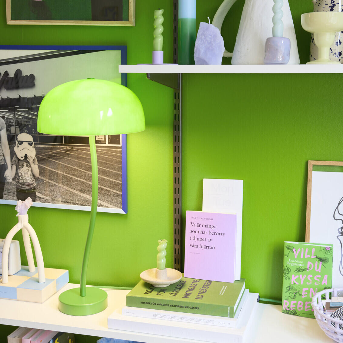 Genevieve sitter vid en limegrön vägg med en Sparring by Elfa bokhylla i vitt och grått med färgstarka prydnadssaker.