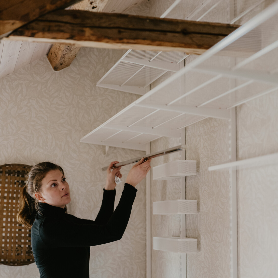 Johanna Berglund asentaa valkoisen Classic by Elfa -säilytyskalusteen ullakolle kattopalkeilla.