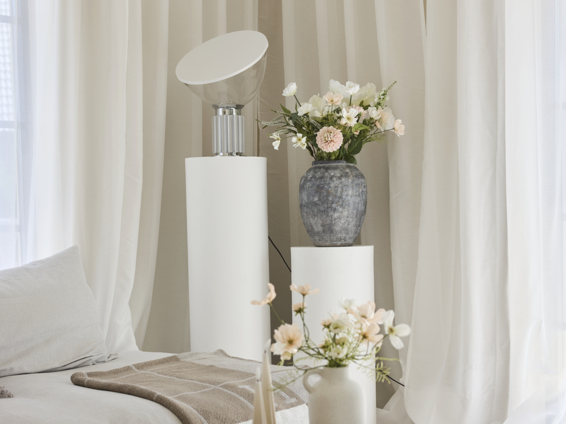 Table lamp and vases with flowers in light colours on a pedestal by a sofa.