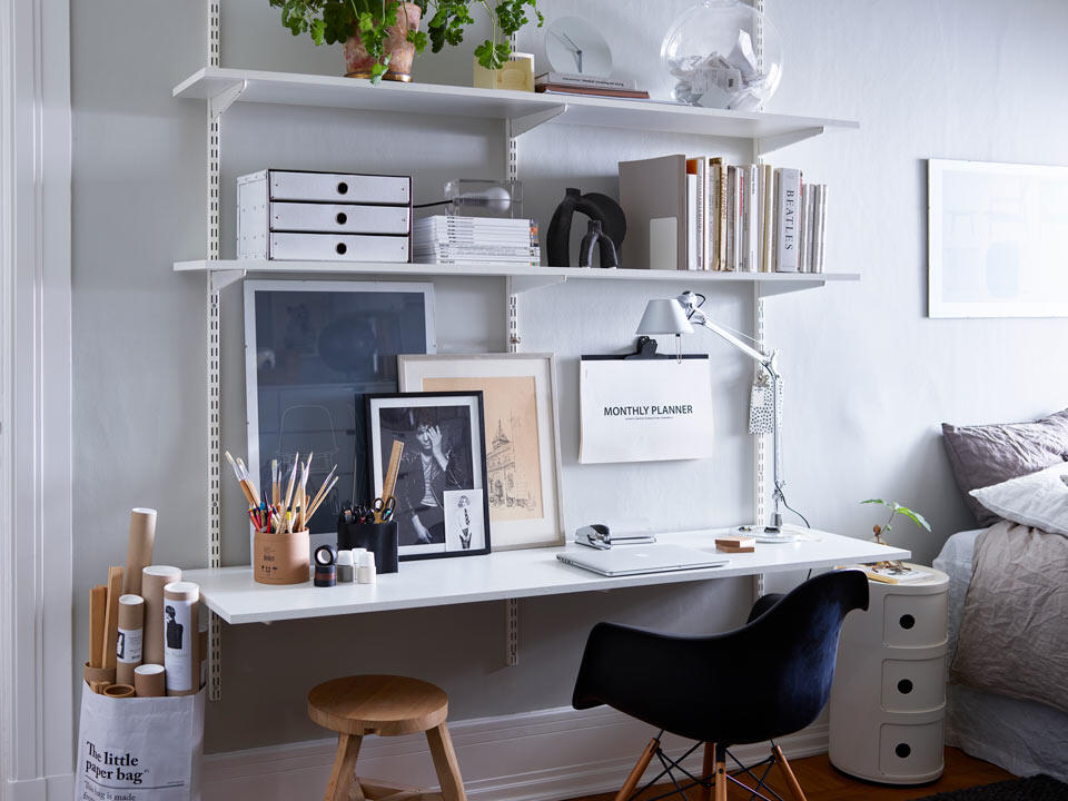 Domowe biuro z klasycznym systemem półek ściennych „Sparring” Elfa, czarnym fotelem biurowym i drewnianym stołkiem. 