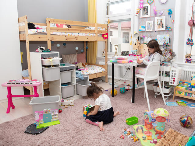 Dětský pokoj Haga s patrovou postelí, stolem a hračkami rozmístěnými na růžovém koberci, potřebuje úložný prostor