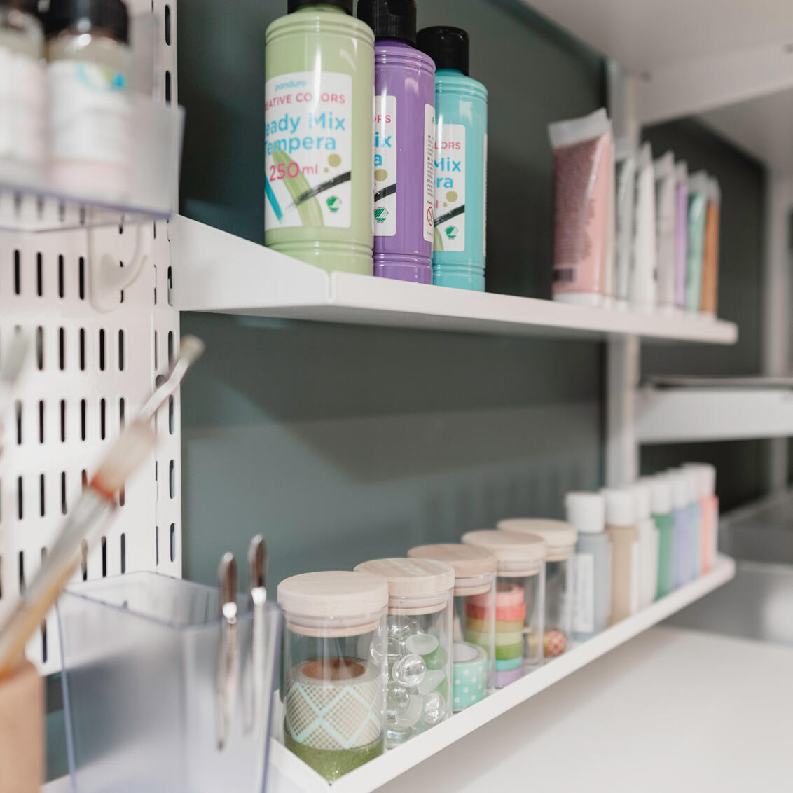 Półki Elfa z uporządkowanymi butelkami z farbami i materiałami rzemieślniczymi