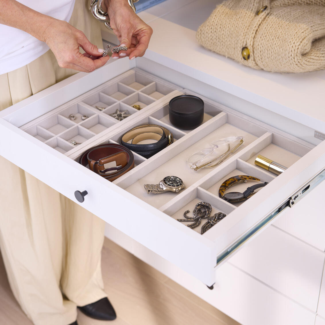  En kvinne står ved en uttrukket Décor+ skuff som inneholder tilbehørsbrett fylt med smykker og annet tilbehør. 