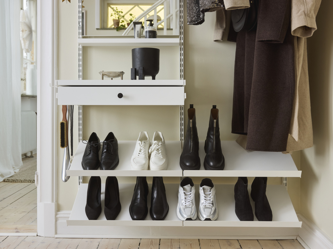 Liten gang med Décor+ by Elfa garderobe i hvitt med hengende klær og skrånende hyller med sko på.