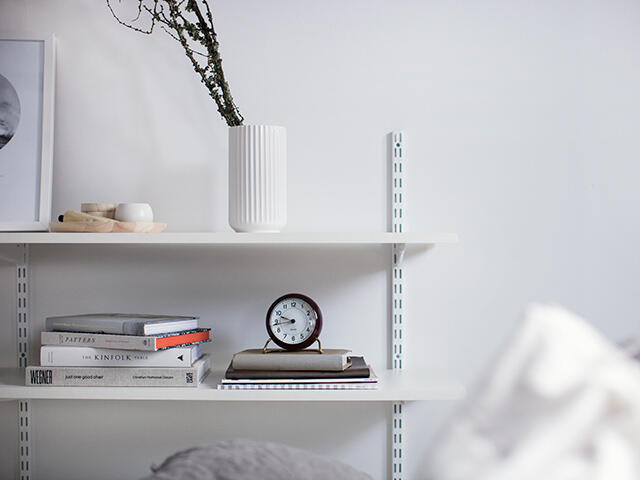 Białe półki Elfa z barierkami ściennymi przedstawiającymi książki, dekoracje i mały zegar