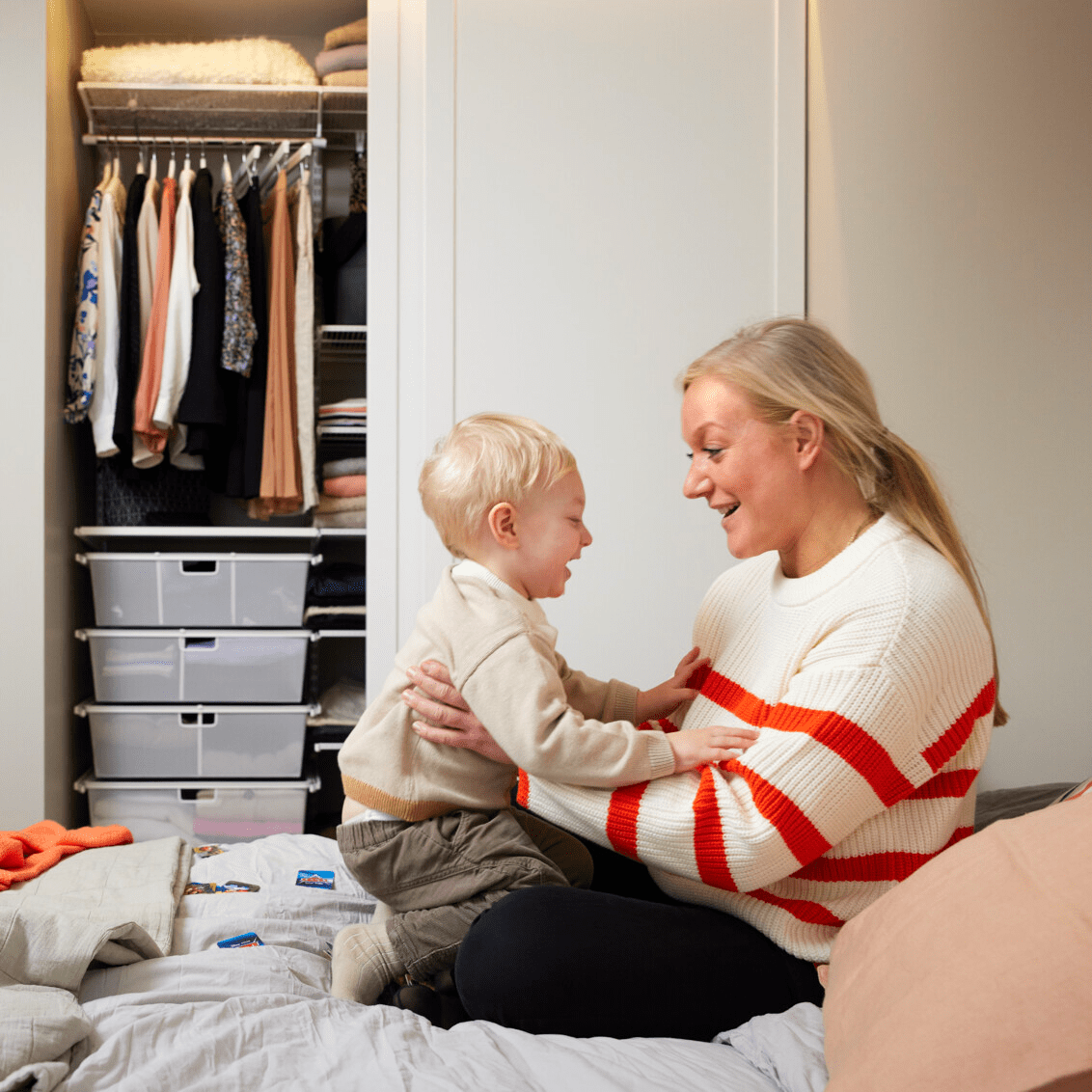 A mother holds her little boy. In the background is an open wardrobe with sliding doors and clothes storage.